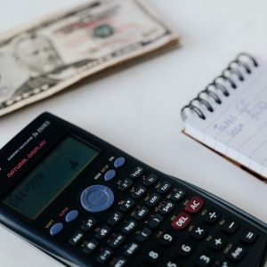 Close-up of a calculator, US dollar bill, and a hand writing in a notebook, symbolizing financial planning.